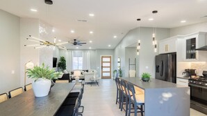 A modern kitchen with a central island, a dining area with a long table, and a living room with a sofa and a TV. - Calypso by Avantstay Entertainers Paradise Pool, Hot Tub, Putting Green (Indio)