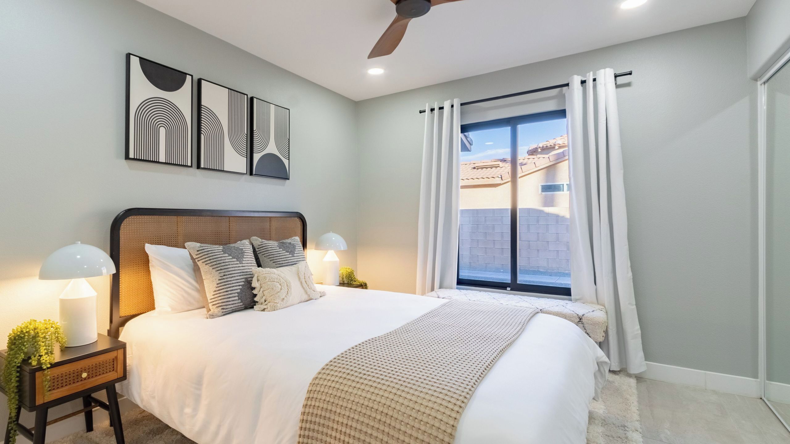 Une chambre à coucher avec un lit, des lampes de chevet, un ventilateur de plafond, une fenêtre avec des rideaux et des œuvres d’art accrochées au mur.