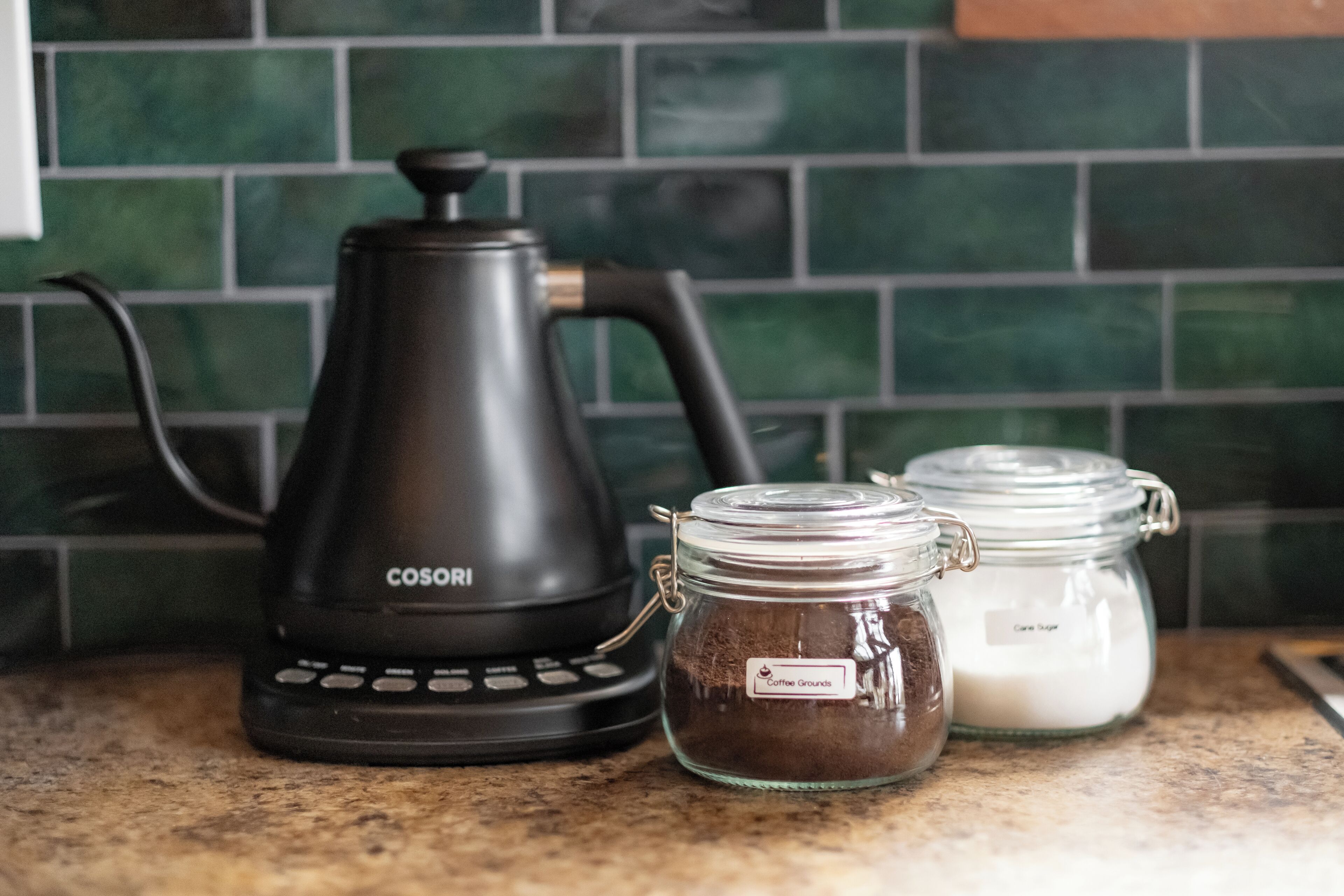 A kitchen with a black coffee maker, two glass jars labeled with coffee grounds and creamer, and a green tiled backsplash.