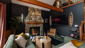 Un salon chaleureux avec une cheminée en pierre, des canapés verts, une table basse ronde et un téléviseur avec Netflix.