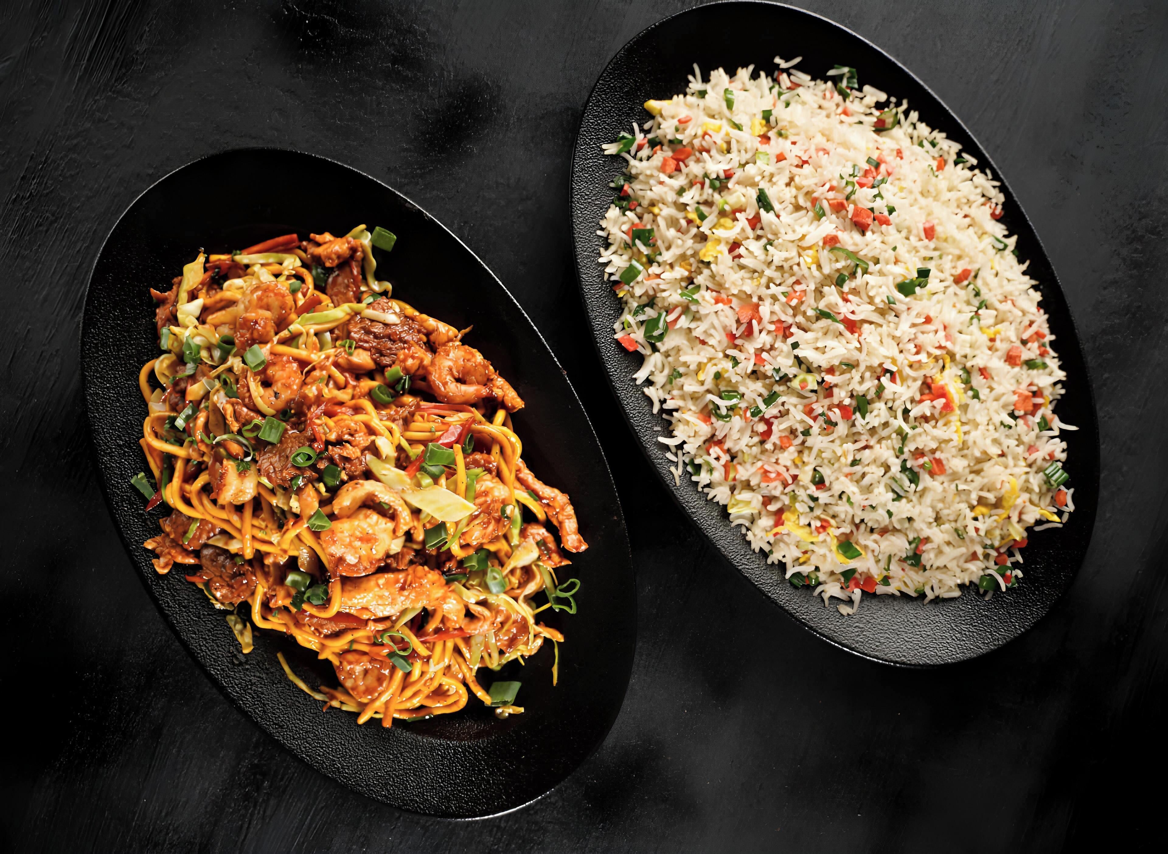Dois pratos, um com macarrão e carne, outro com arroz e legumes, sobre uma superfície escura.