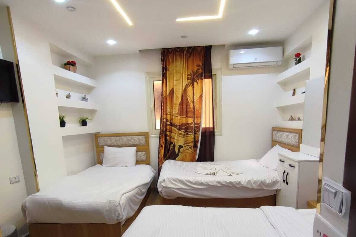 A hotel room with two beds, a ceiling with geometric lighting, a curtain with a desert scene, and built-in shelves with items.