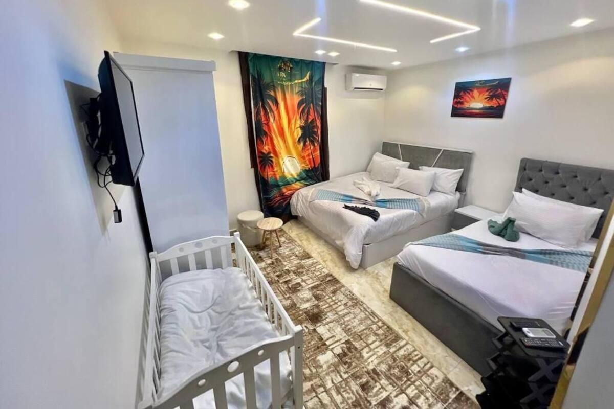 A hotel room with a crib, two beds, a flat-screen TV, a wall-mounted air conditioner, and a tropical-themed tapestry.