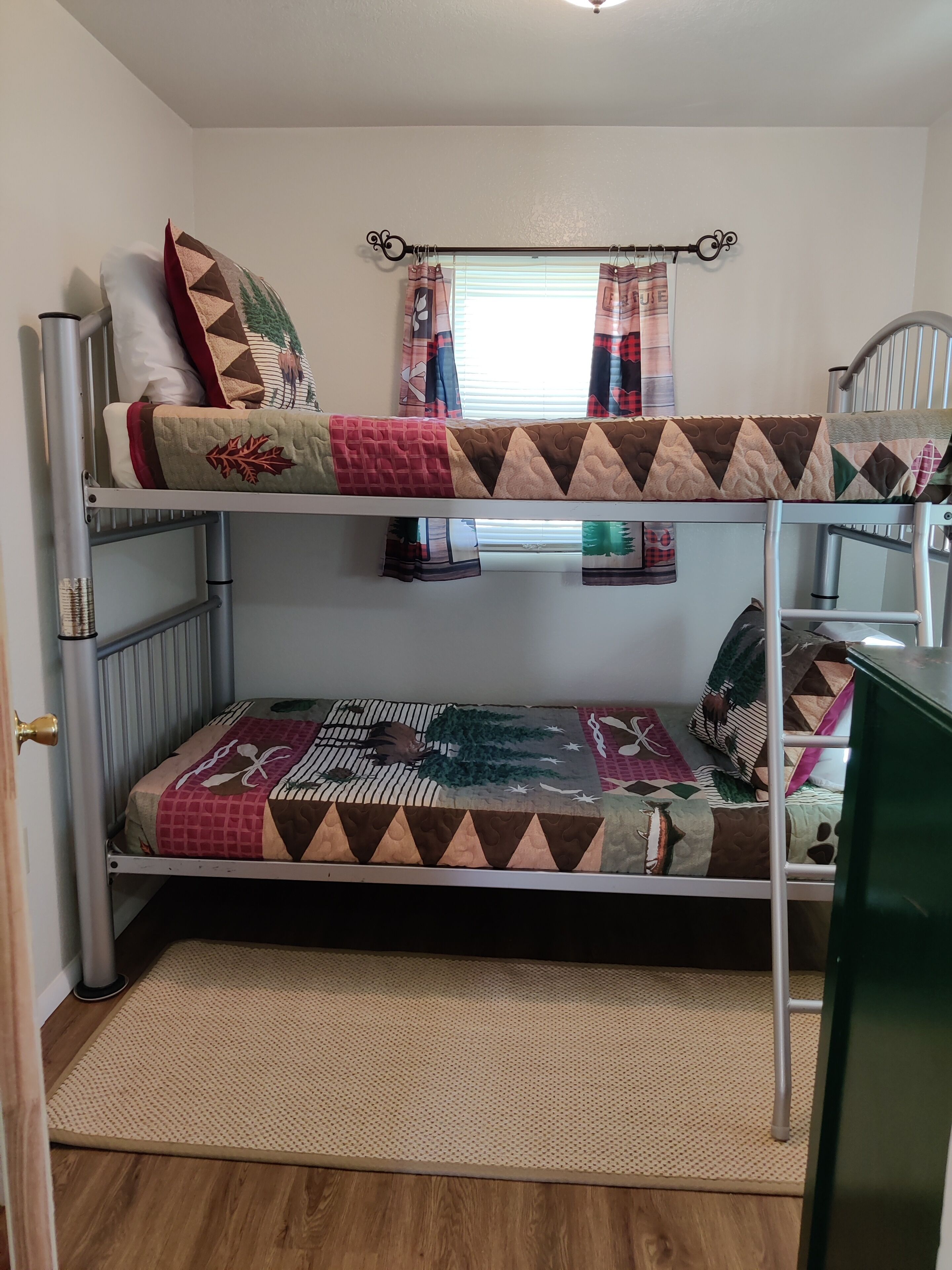 A bunk bed with two levels, each with a quilted bedspread. There is a window with curtains, a radiator, and a door with a brass handle.