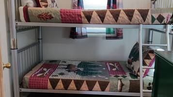 A bunk bed with two levels, each with a quilted bedspread. There is a window with curtains, a radiator, and a door with a brass handle.