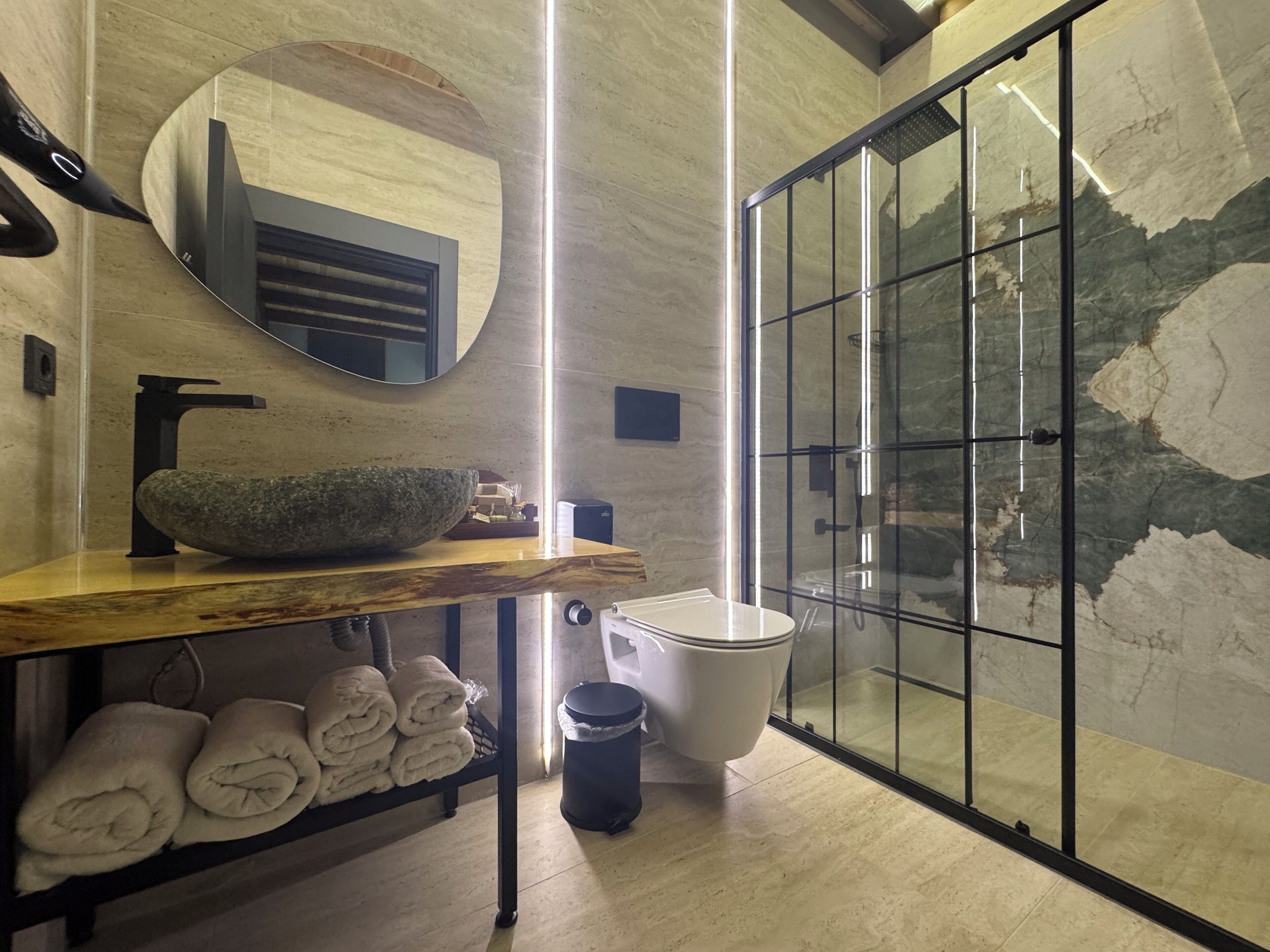 A modern bathroom with a stone basin, a large mirror, a glass shower enclosure, and neatly folded towels on a shelf.