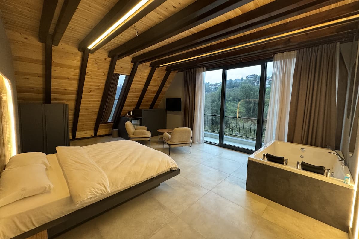 A modern bedroom with a large bed, a sitting area with chairs, a bathtub, and a balcony with a view of the city.