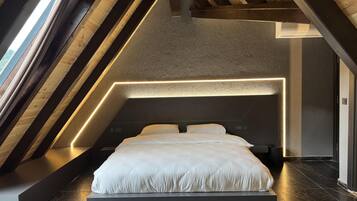 Une chambre de style loft moderne, dotée d’un plafond haut, de poutres apparentes et d’un lit placé au centre, sur un plancher foncé.