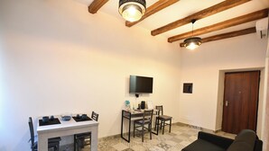 A living room with a sofa, a dining table with chairs, a TV mounted on the wall, and a wooden ceiling beam. - GreSi's home - Holiday home, in the heart of medieval Tivoli (Tivoli)
