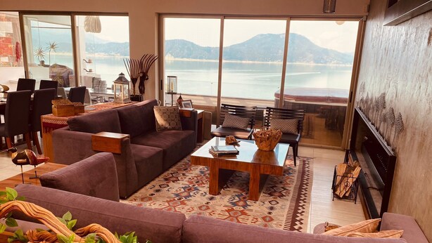 Ein geräumiges Wohnzimmer mit einer großen Couch, einem Couchtisch und einem Kamin. Große Fenster bieten Blick auf das Meer und die Berge.