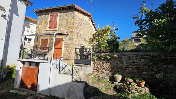Un bâtiment en pierre avec une porte en bois et des volets aux fenêtres, un escalier en métal et des plantes en pot.