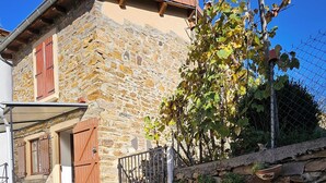 Une maison en pierre avec un toit recouvert de tuiles rouges, une entrée fermée par un portail et un petit jardin agrémenté de plantes en pot.
