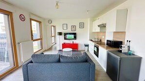 A modern apartment with a grey sofa, a kitchen with a microwave and toaster, a TV on a stand, and a window with a view of the outside.