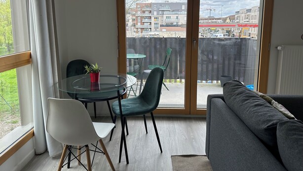 A modern living room with a glass door leading to a balcony, a round glass table with chairs, and a grey sofa.