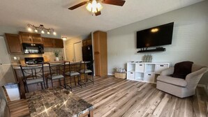 Eine moderne Küche mit Holzausstattung, ein Essbereich mit Marmortisch und ein Wohnzimmer mit Fernseher und Sitzmöbeln.