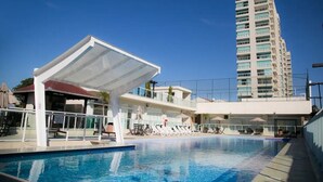 Una piscina con sillas de descanso y un área de estar techada, rodeada por un edificio alto.