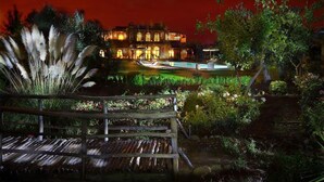 A well-lit garden with a wooden deck, a pool, and a large building in the background.
