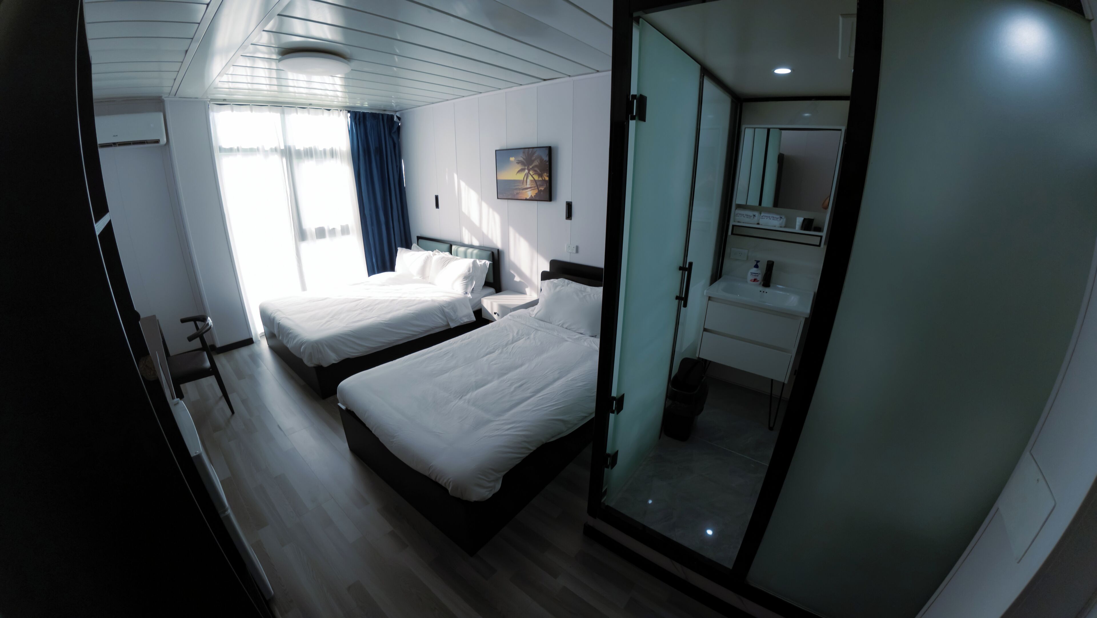 Una habitación de hotel con dos camas, un baño con lavabo y espejo, y un ventanal grande con cortinas.