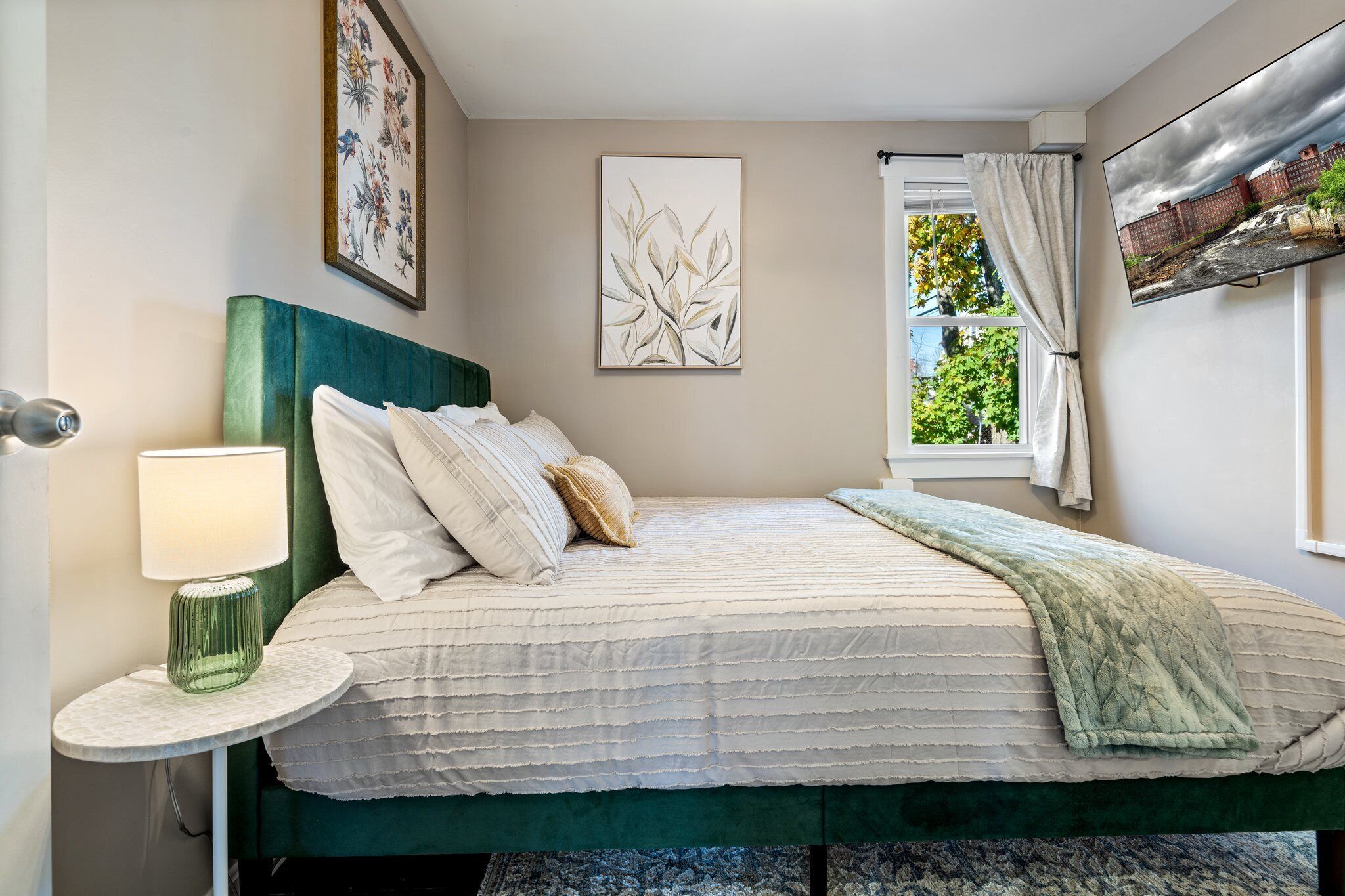 Un dormitorio con cama, una mesita de noche con lámpara, una ventana con cortinas y obras de arte en las paredes.