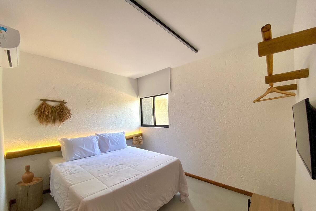 Une chambre à coucher avec un lit, une tête de lit en bois, une petite table de chevet, un téléviseur fixé au mur et une fenêtre.