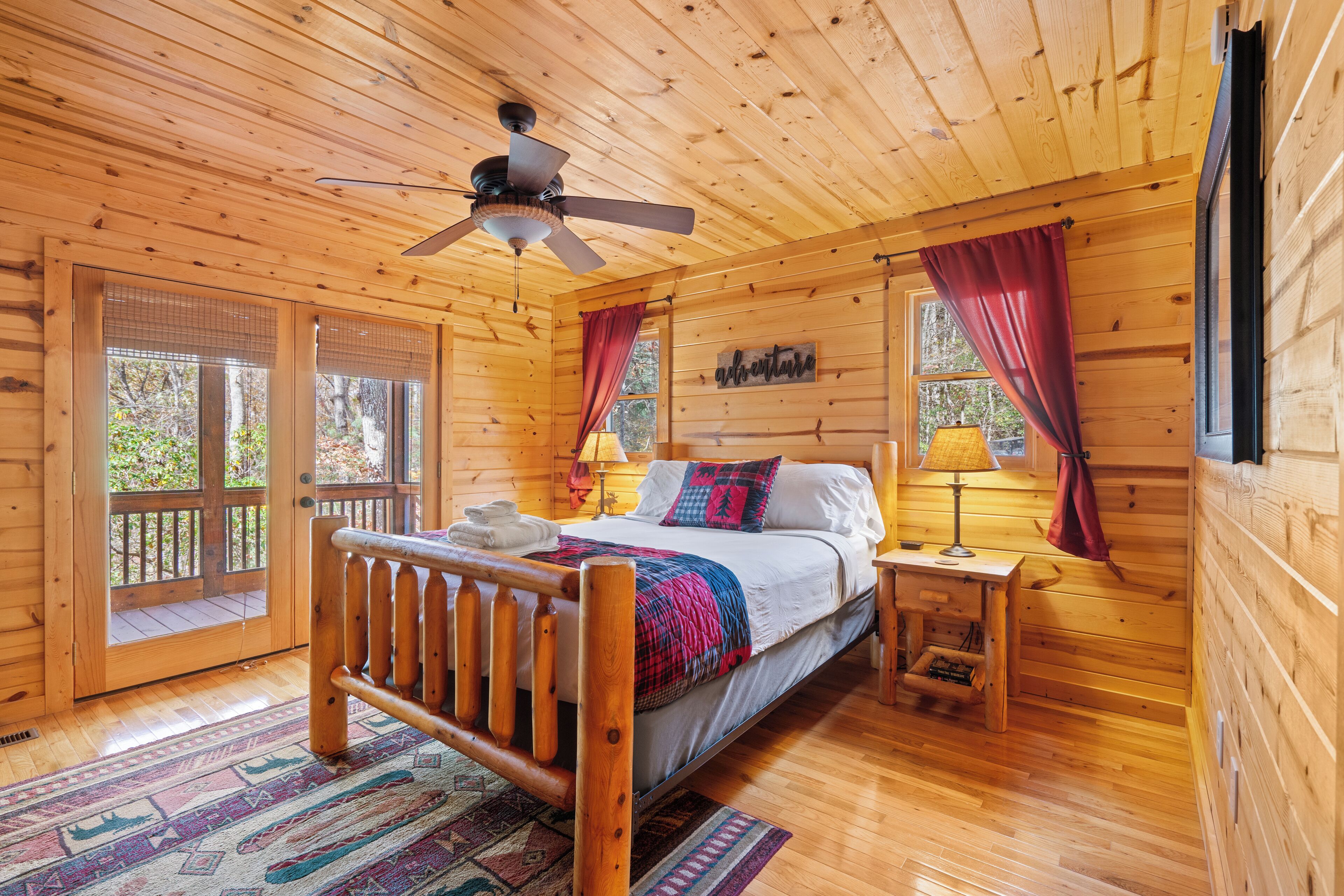 A wooden cabin bedroom with a bed, a nightstand, a ceiling fan, and a window with curtains.