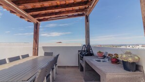 Un patio en la azotea con una pérgola de madera, una mesa de comedor con sillas, una pequeña cocina y plantas en macetas.