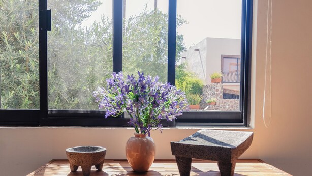 Una mesa de madera con un jarrón de flores moradas, un cuenco pequeño y una escultura de piedra junto a una ventana.