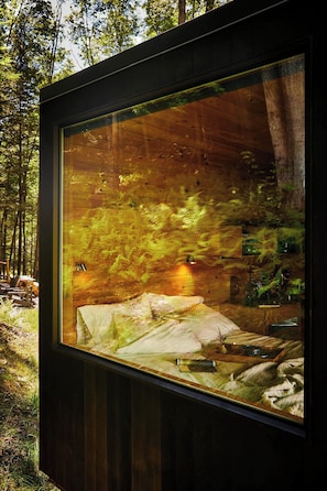 Een glazen omheining met uitzicht op een bos, inclusief een bed en een klein tafeltje.
