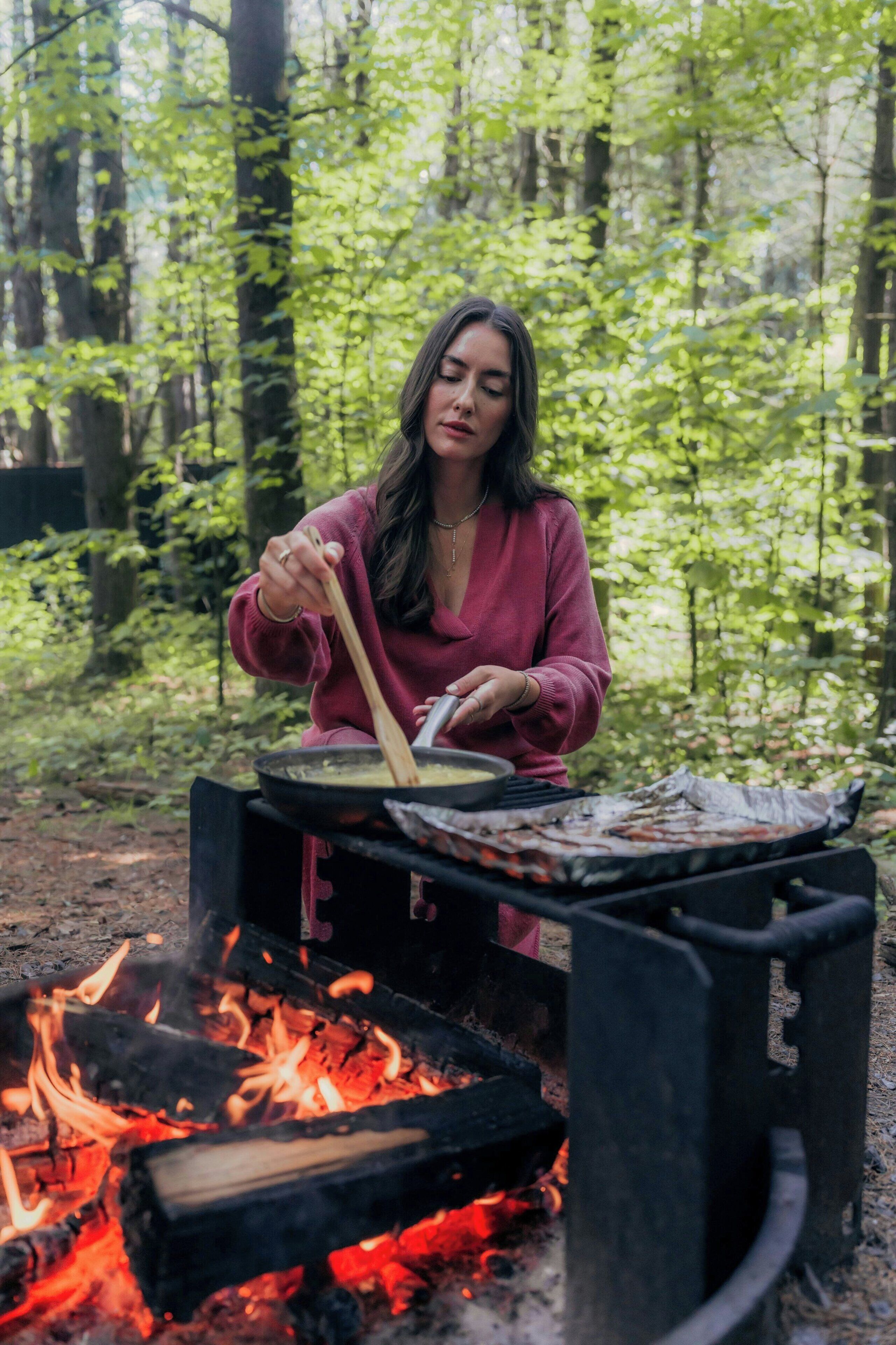 Eine Person kocht draußen mit einem Campingkocher über offenem Feuer.
