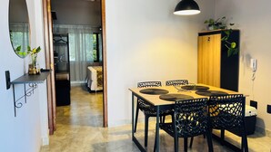 A dining area with a wooden table, black chairs, and a hanging pendant light. A mirror and a shelf with plants are on the wall. - Flat Itajuru (Cabo Frio)