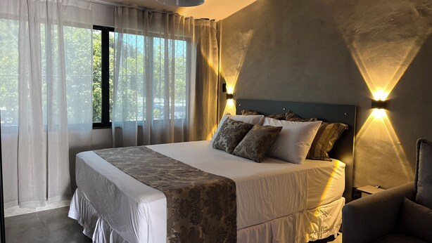 A modern bedroom with a large bed, a headboard with integrated lighting, sheer curtains, and a view of greenery outside. - Flat Itajuru (Cabo Frio)