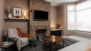 A cozy living room with a stone fireplace, a flat-screen TV mounted above it, a comfortable sofa, a small round coffee table, and a bookshelf with decorative items.