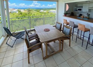 A balcony with a wooden table and chairs, a view of a green landscape, and a kitchen area with a microwave and cabinets.