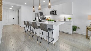 Una cocina moderna con una isla central, taburetes de bar, luces colgantes y armarios blancos.