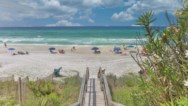 Una playa con un paseo marítimo de madera que lleva a la orilla, rodeada de vegetación y sombrillas de playa.