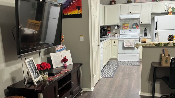 Una cocina moderna con armarios blancos, una estufa y un refrigerador. Hay un comedor con una mesa y sillas, y un televisor montado en la pared.