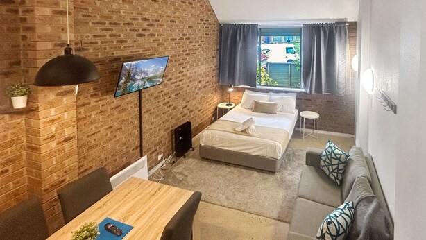 Habitació acollidora amb paret de maó, un llit, un sofà, una taula i una finestra amb cortines.