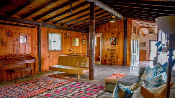 Interior rústico de una cabaña con un piano de madera, un sofá de cuero, un tapete estampado y un diván con almohadones.
