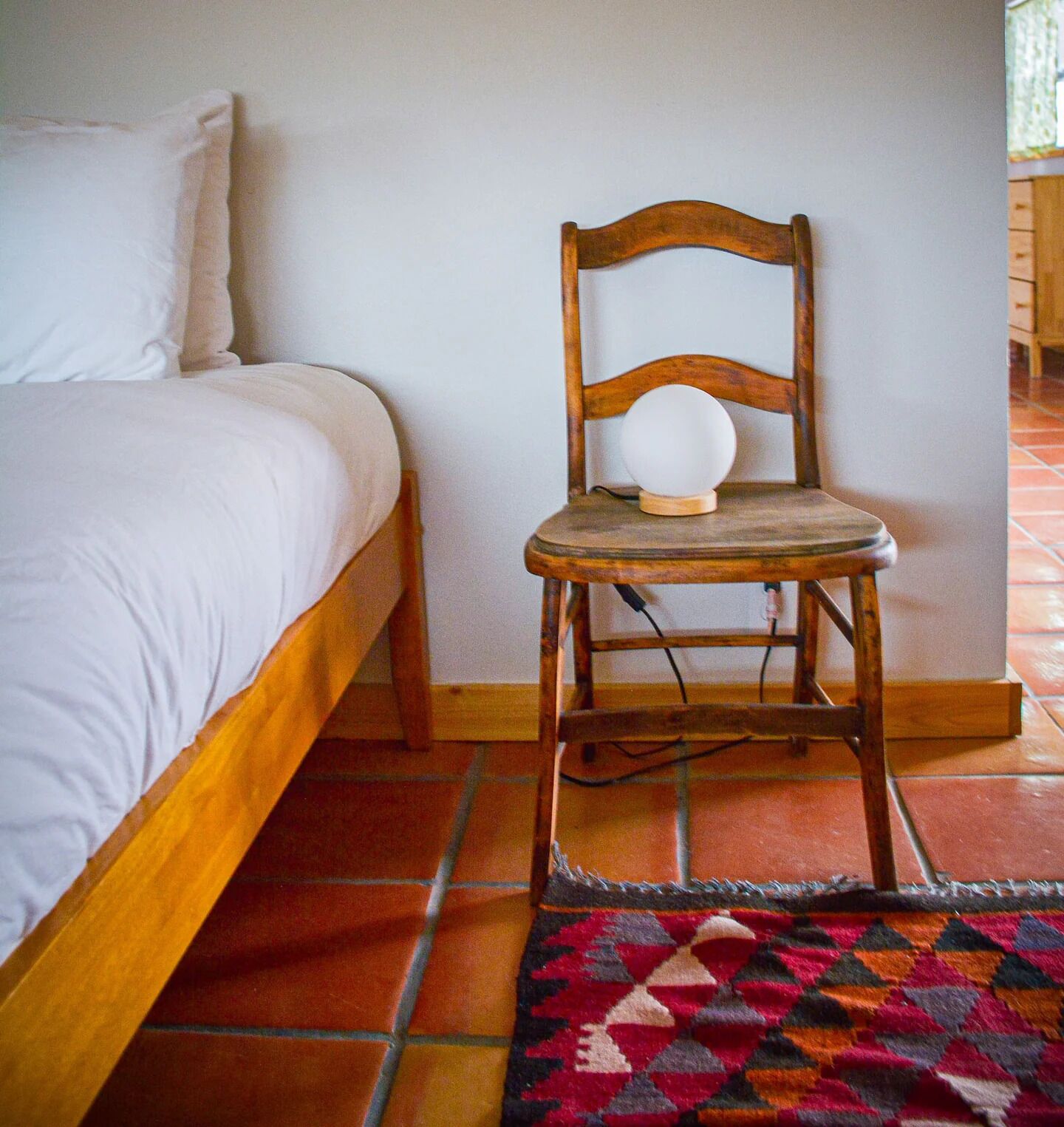 Une chambre avec un lit en bois, une chaise en bois sur laquelle est posé un objet blanc, et un tapis à motifs sur le sol.