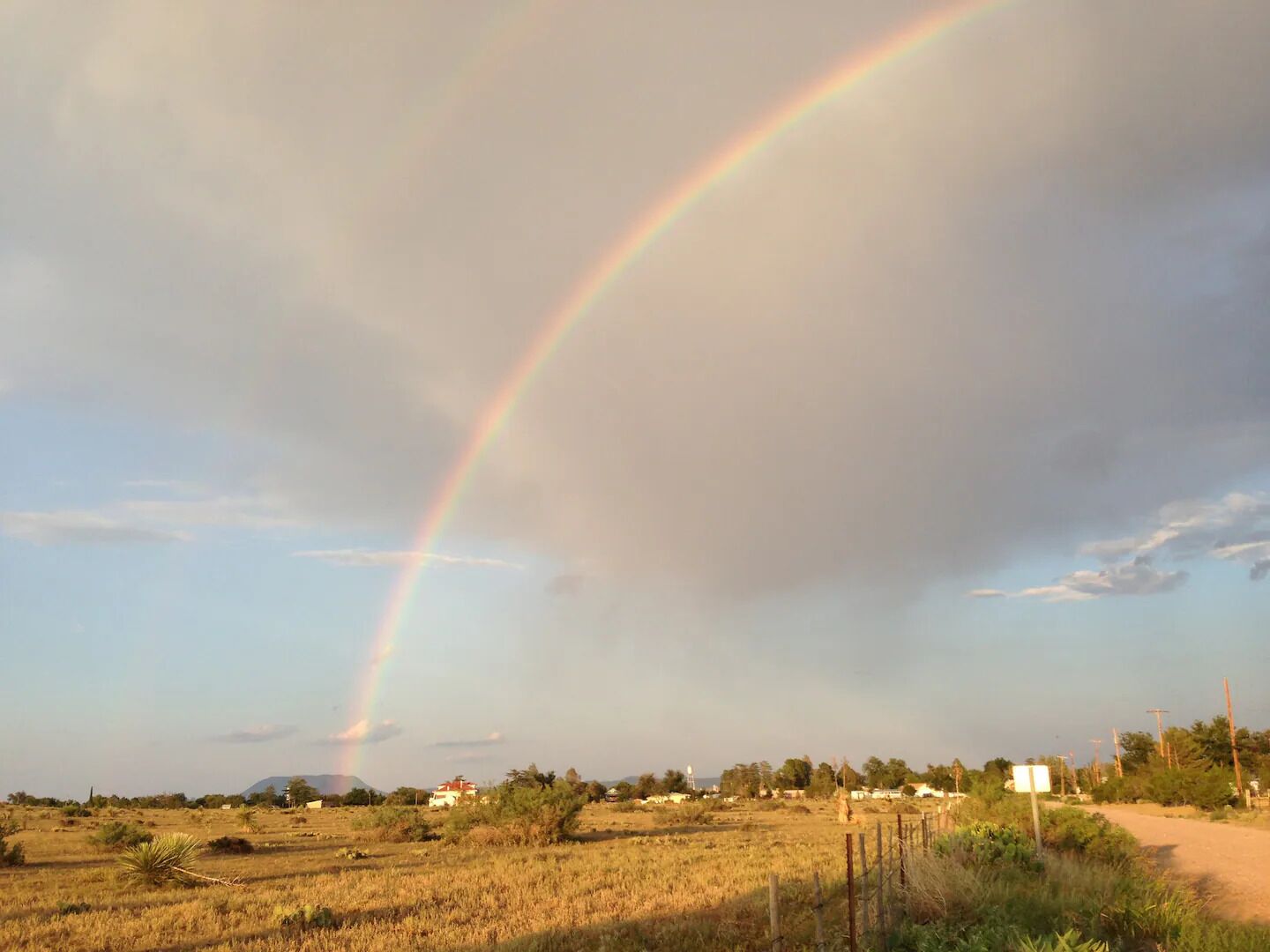 Paesaggio rurale con un arcobaleno, campi aperti, alcuni edifici e una strada sterrata.