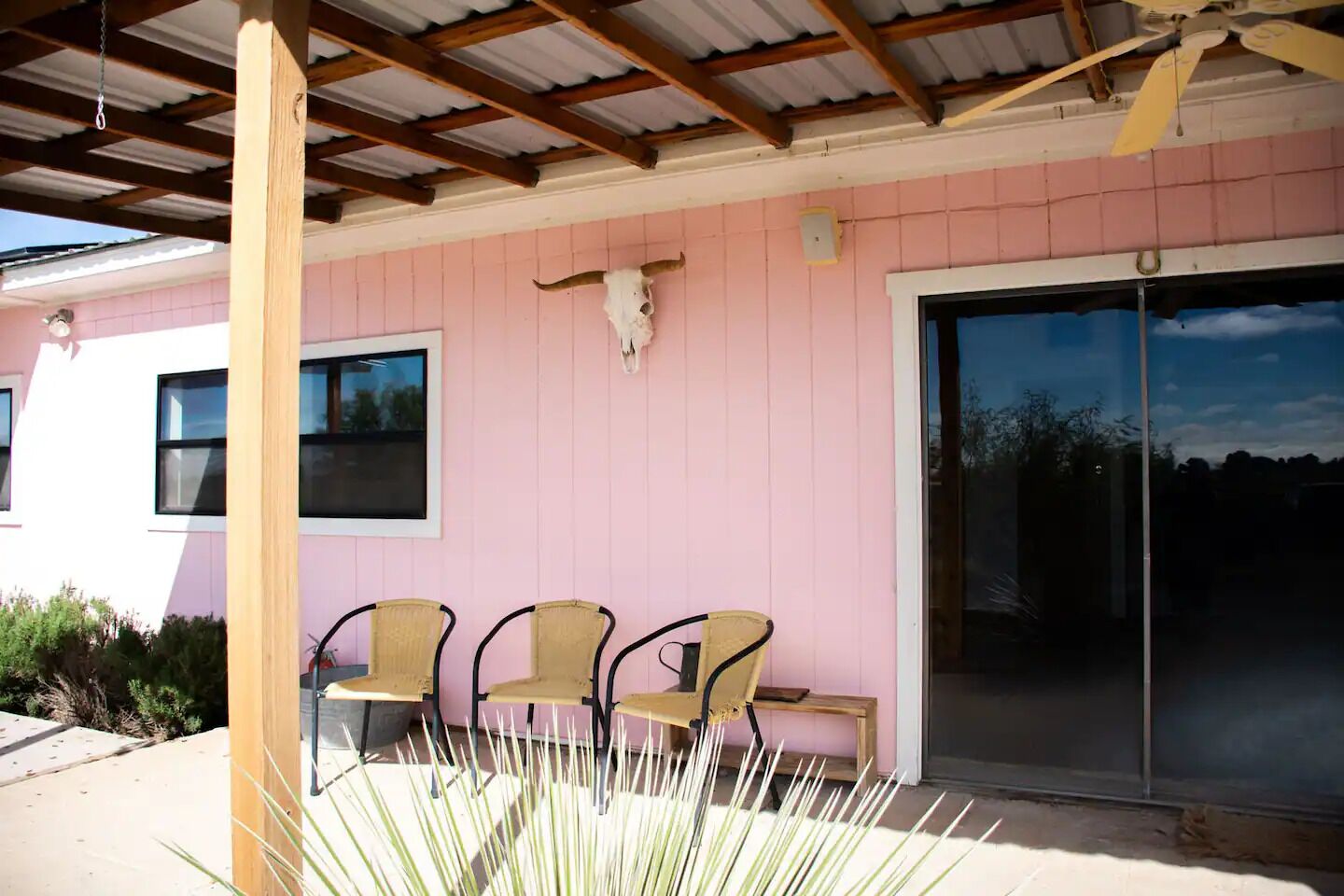 A covered patio with three chairs, a wooden post, and a large glass door.