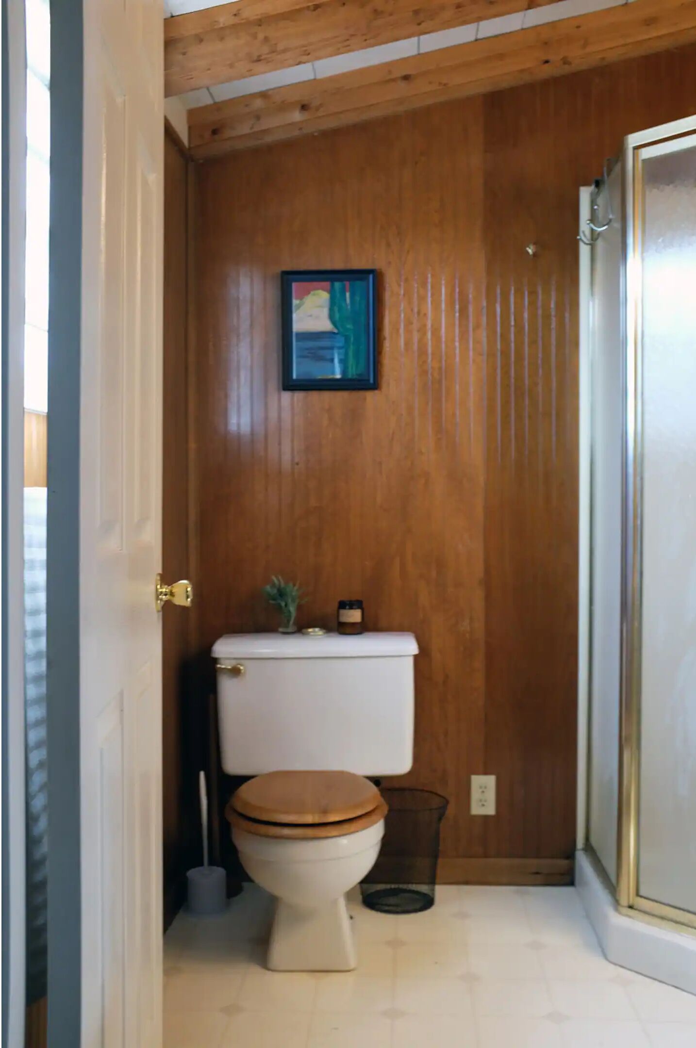 A small bathroom with wooden walls, a white toilet, a wooden seat, a small potted plant, and a trash can.