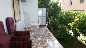 Un patio con mobiliario de mimbre y suelo de baldosas, rodeado de follaje y un edificio al fondo.