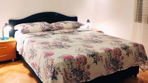 Un dormitorio con piso de madera, una cama con cubrecama floral, una mesita de noche con lámpara y un equipo de aire acondicionado en la pared.