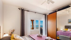 A hotel room with a bed, a window with a view of the sea, a ceiling fan, and a wooden door.