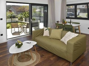 A modern living room with a green sofa, a small white table, and a view of an outdoor patio with chairs and a table.