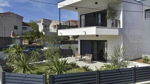 A modern two-story house with a balcony, a patio with outdoor furniture, and a fenced garden area.