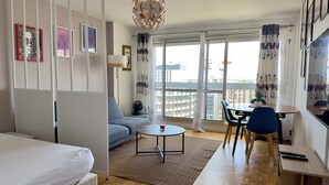 A modern hotel room with a bed, a sofa, a dining area with a table and chairs, a small rug, and a view of the city through large windows. - Spacious 2-room apartment with parking in Paris La Défense overlooking the Eiffel Tower   (Puteaux)