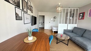 A modern living room with a wooden dining table, a grey sofa, and a blue chair. There are framed pictures on the wall and a chandelier. - Spacious 2-room apartment with parking in Paris La Défense overlooking the Eiffel Tower   (Puteaux)