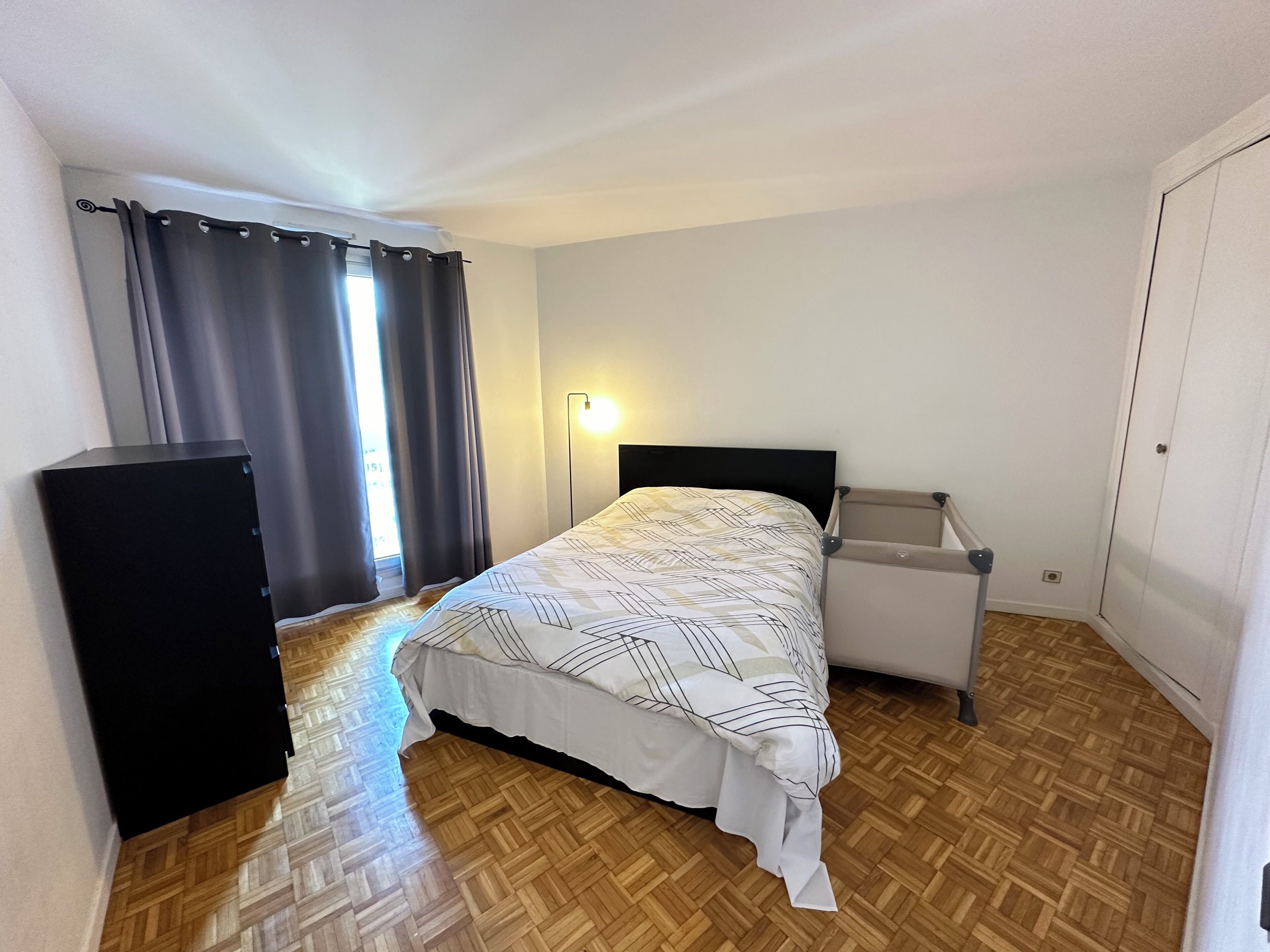 A bedroom with a bed, a bedside table, a lamp, a wardrobe, and a window with curtains.